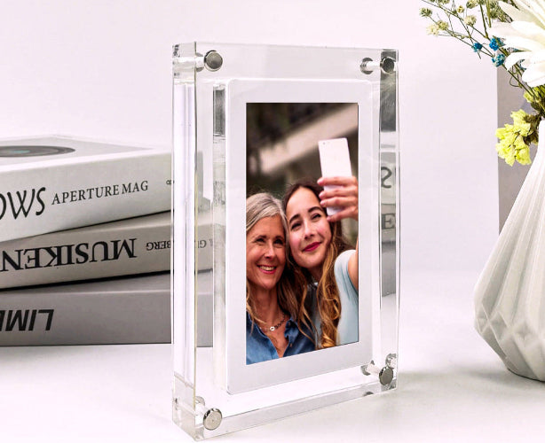 Clear glass photo frame with a photo of two women on a white surface with books and a vase in the background.
